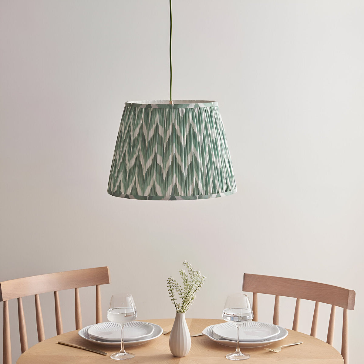 Brackmont Pendant Light with pleated green fabric shade and brass flex, hanging over a wooden dining table in a light-toned room.