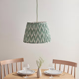 Brackmont Pendant Light with pleated green fabric shade and brass flex, hanging over a wooden dining table in a light-toned room.