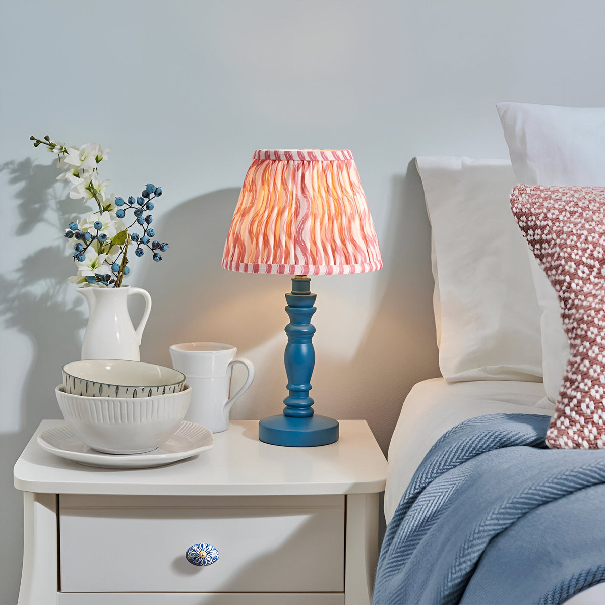 Endon Aldworth Ripple table lamp with blue turned spindle base and pleated coral pink ripple-print shade on a white bedside table beside a bed.