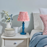 Endon Aldworth Ripple table lamp with blue turned spindle base and coral pink pleated ripple-print shade on a white bedside table beside a bed.