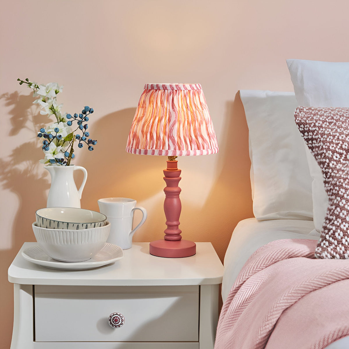 Aldworth Ripple Table Lamp with pink turned spindle base and coral ripple-print fabric shade, styled on a bedside table beside a bed.