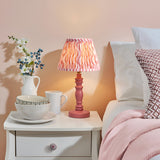Aldworth Ripple Table Lamp with pink turned spindle base and coral ripple-print fabric shade, styled on a bedside table beside a bed.