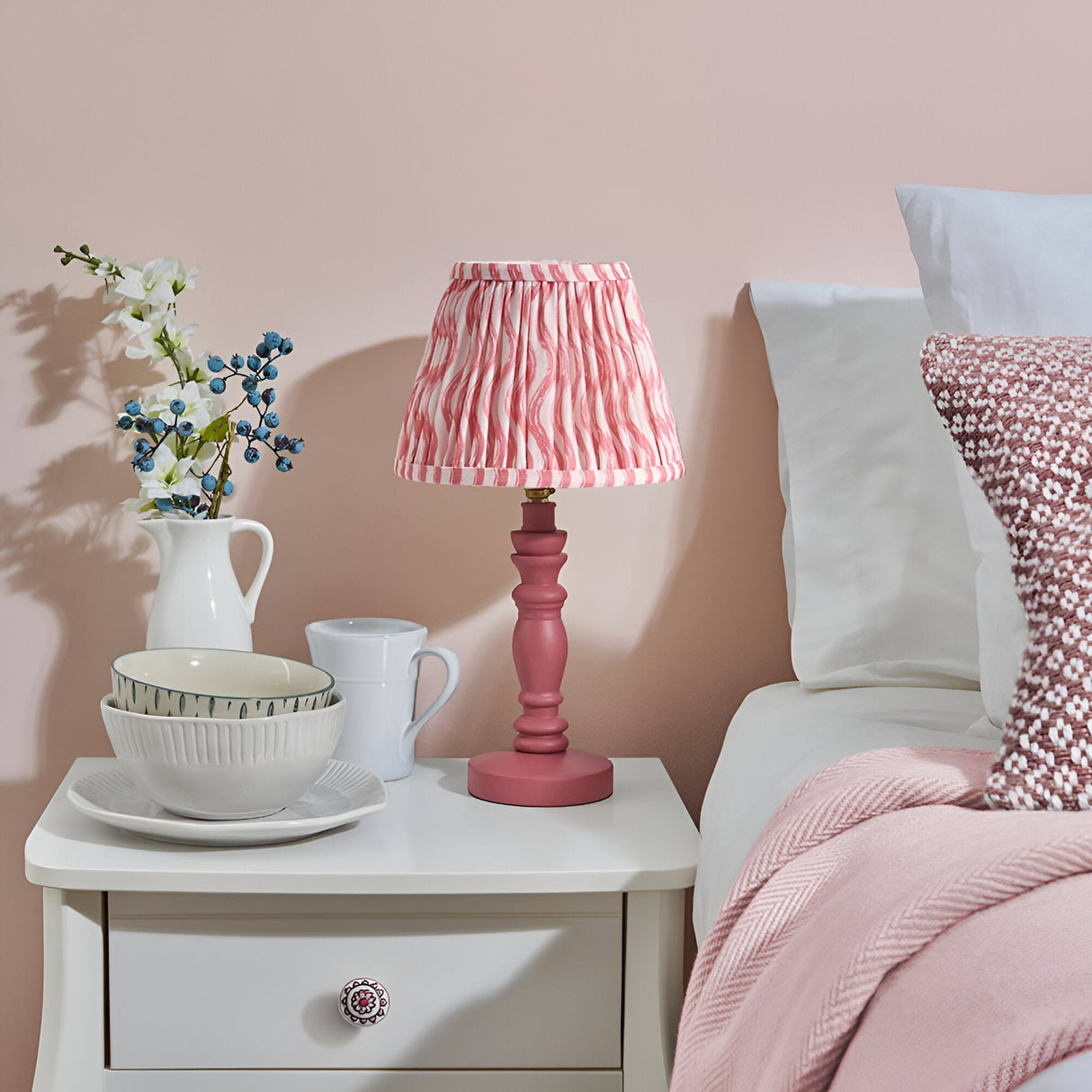 Aldworth Ripple Table Lamp with pink spindle base and pleated pink ripple-print fabric shade, styled on a bedside table beside pink bedding.