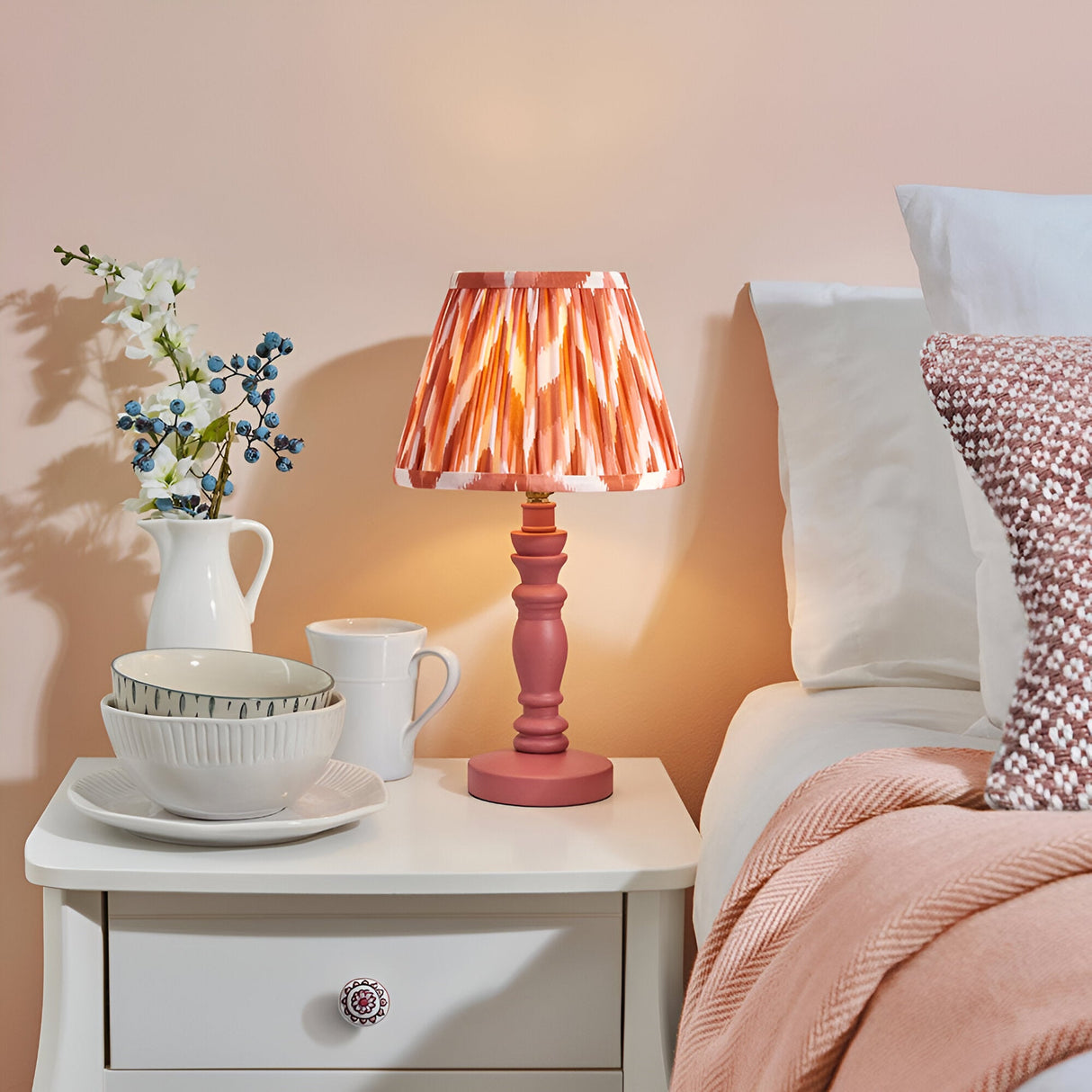Aldworth Zigzag Table Lamp with pink patterned shade and turned wooden base, shown lit on a white bedside table beside a bed.