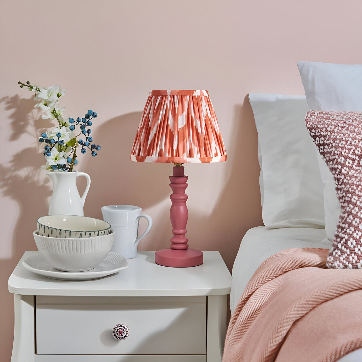 Aldworth Zigzag Table Lamp with pink turned wood base and pink zigzag pleated shade, styled on a white bedside table beside a jug of flowers and bedding.