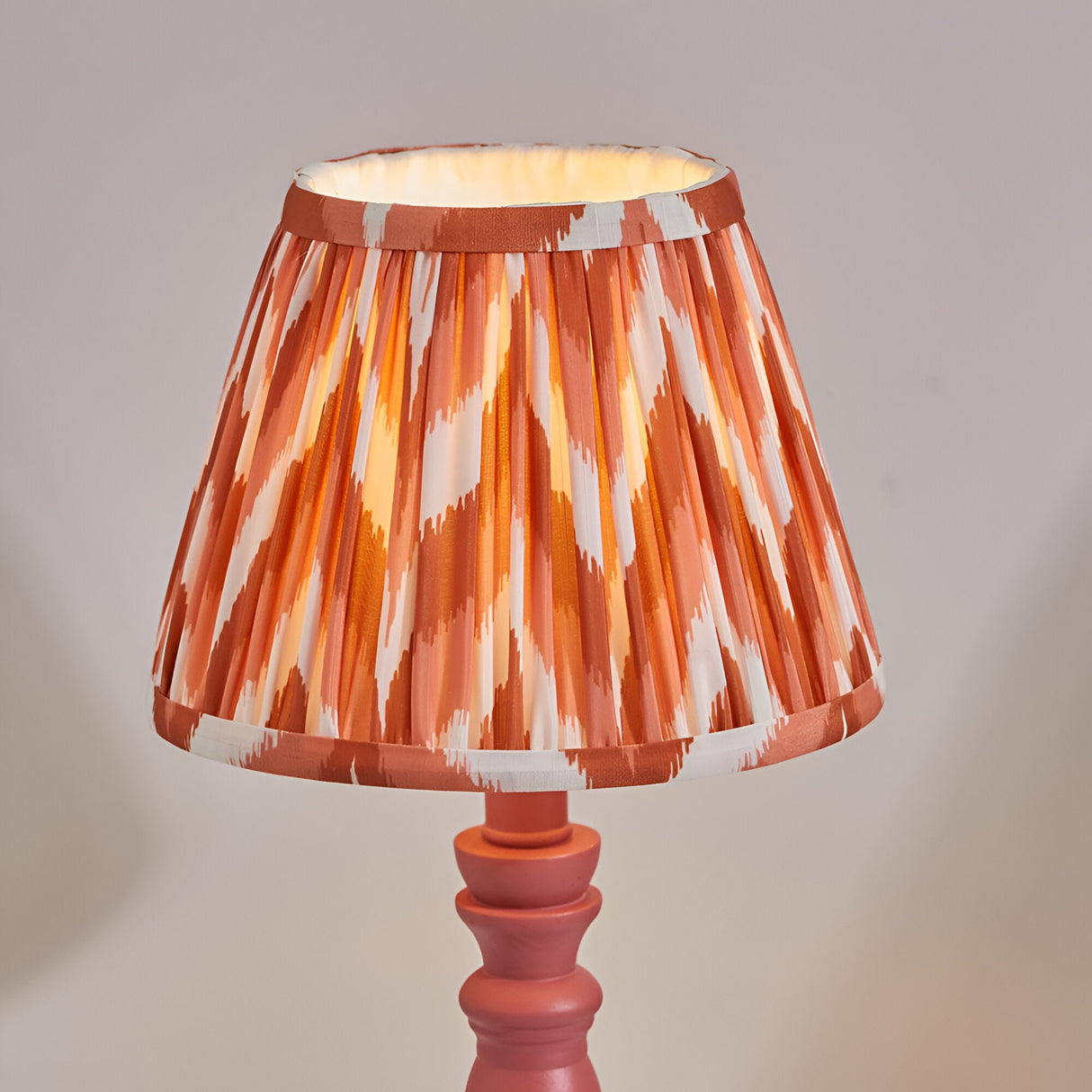 Aldworth table lamp with pink turned wood base and pleated zigzag-print fabric shade in orange, white and rust tones, on a plain light background.