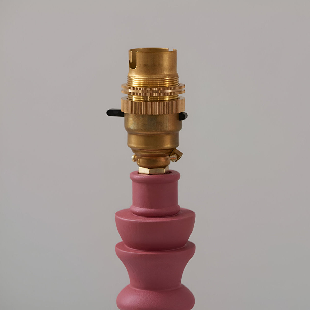 Aldworth Zigzag Table Lamp base close-up showing a brass B22 switched lamp holder atop a hand-turned pink wooden spindle base, plain grey background.