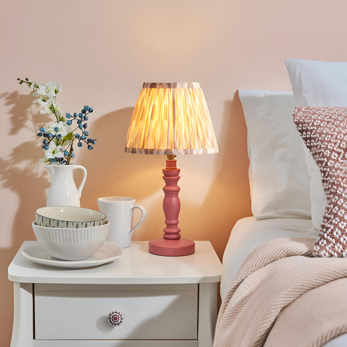 Endon Aldworth Ikat table lamp with pink turned spindle base and pleated ikat shade, lit on a white bedside table beside a bed with soft pink decor.
