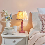 Endon Aldworth Ikat table lamp with pink turned spindle base and pleated ikat shade, lit on a white bedside table beside a bed with soft pink decor.