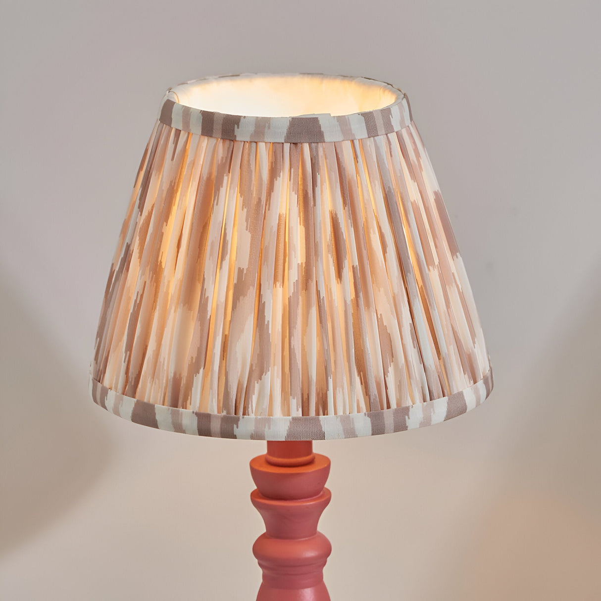 Endon Aldworth Ikat table lamp with pleated natural and grey ikat print shade on a pink turned wooden spindle base, illuminated against a neutral background.