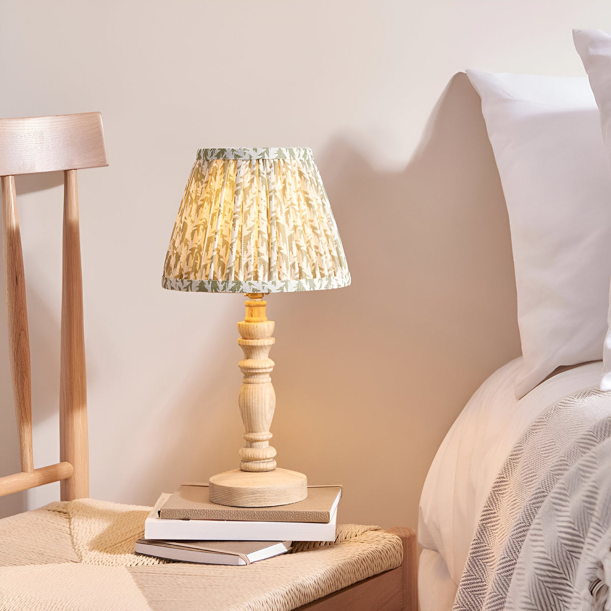 Aldworth Leaf Table Lamp in natural ash with a pleated leaf-print shade, shown on a stack of books beside a bed in a bedroom setting.