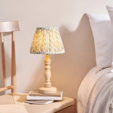 Aldworth Leaf Table Lamp in natural ash with a pleated leaf-print shade, shown on a stack of books beside a bed in a bedroom setting.