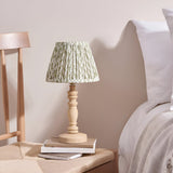 Aldworth Leaf table lamp with turned natural ash spindle base and pleated green leaf-print shade, styled on bedside books beside a bed.