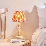 Endon Aldworth table lamp with turned natural wood spindle base and pleated zigzag shade in orange tones, on books beside a bed.