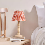 Endon Aldworth table lamp with turned natural wood base and pleated coral zigzag faux silk shade, on books beside a bed with white linens.