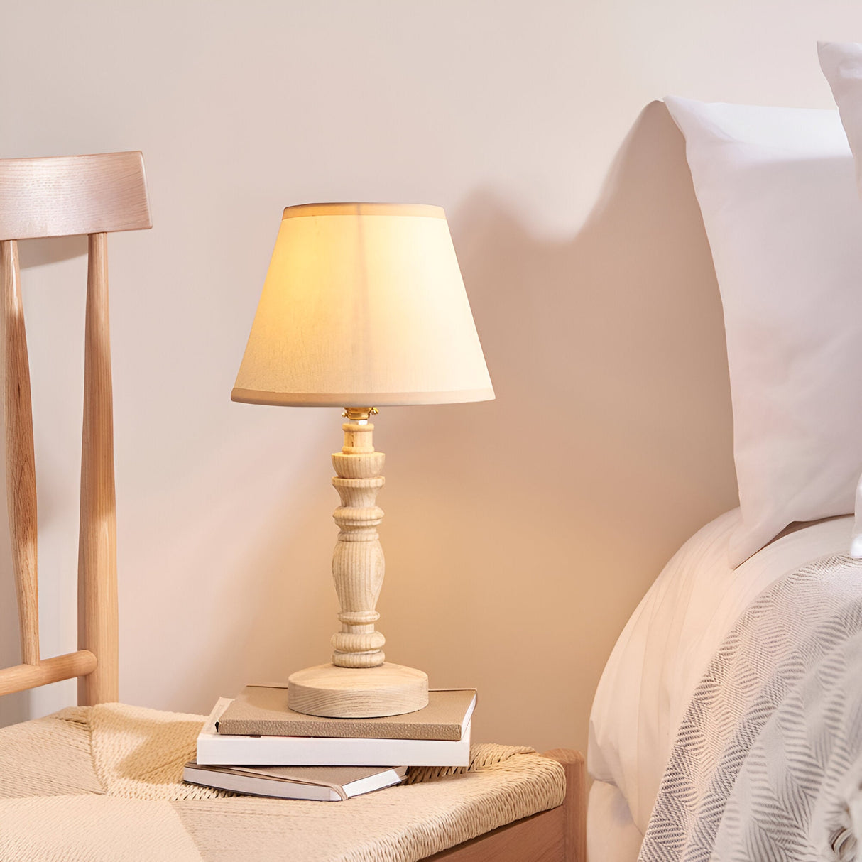 Endon Aldworth table lamp with turned natural ash spindle base and tapered vintage white shade, lit on a bedside stack of books beside a bed.
