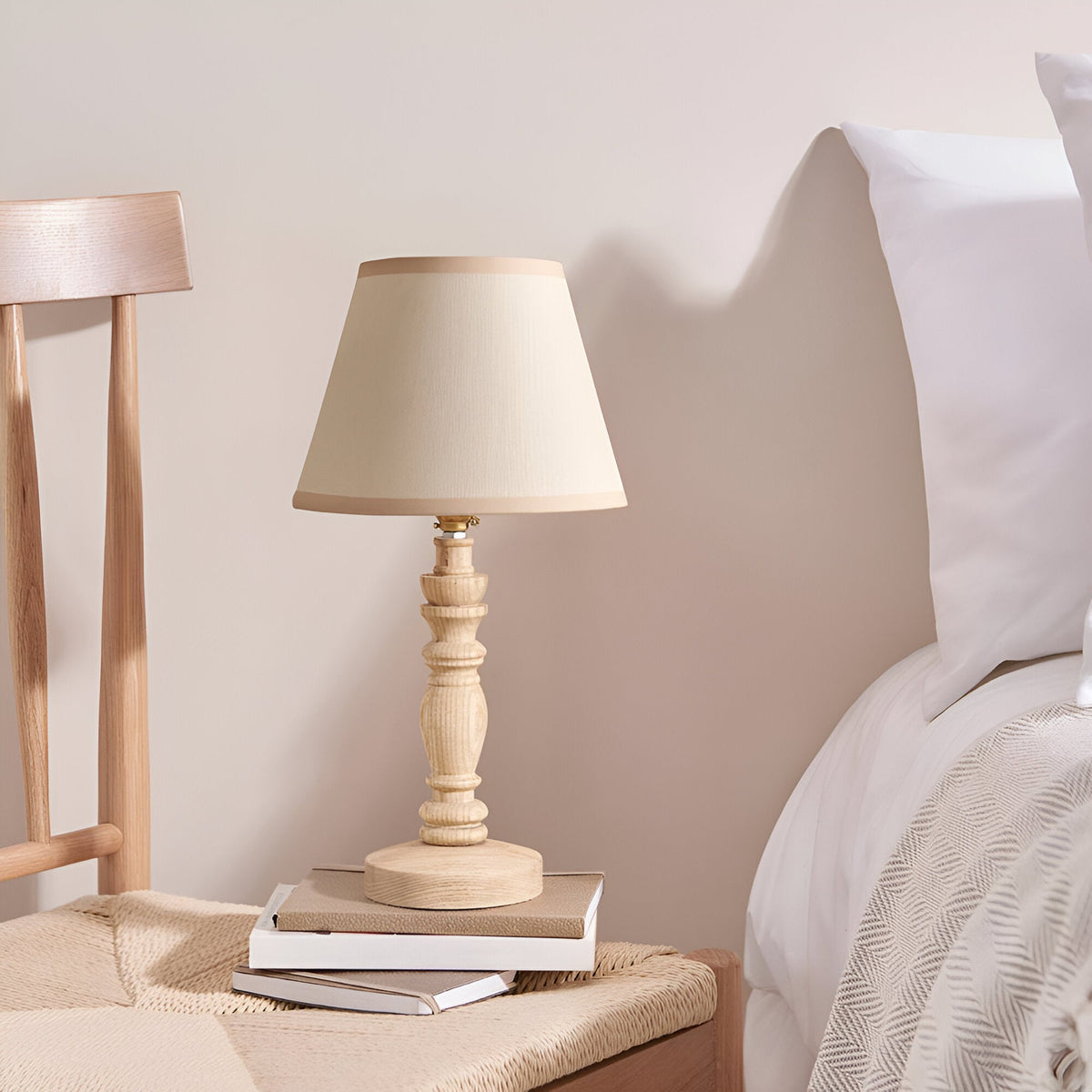 Endon Aldworth table lamp with turned natural ash spindle base and tapered vintage white shade on a stack of books beside a bed.