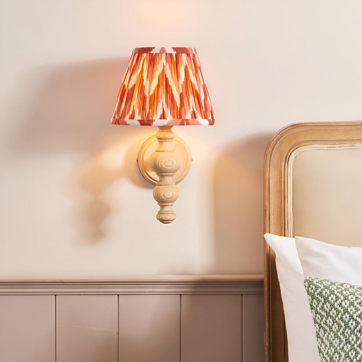 Aldworth Zigzag Wall Light in natural ash with turned spindle base and pleated apricot orange zigzag shade, shown lit above a bedroom headboard.
