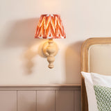 Aldworth Zigzag Wall Light in natural ash with turned spindle base and pleated apricot orange zigzag shade, shown lit above a bedroom headboard.