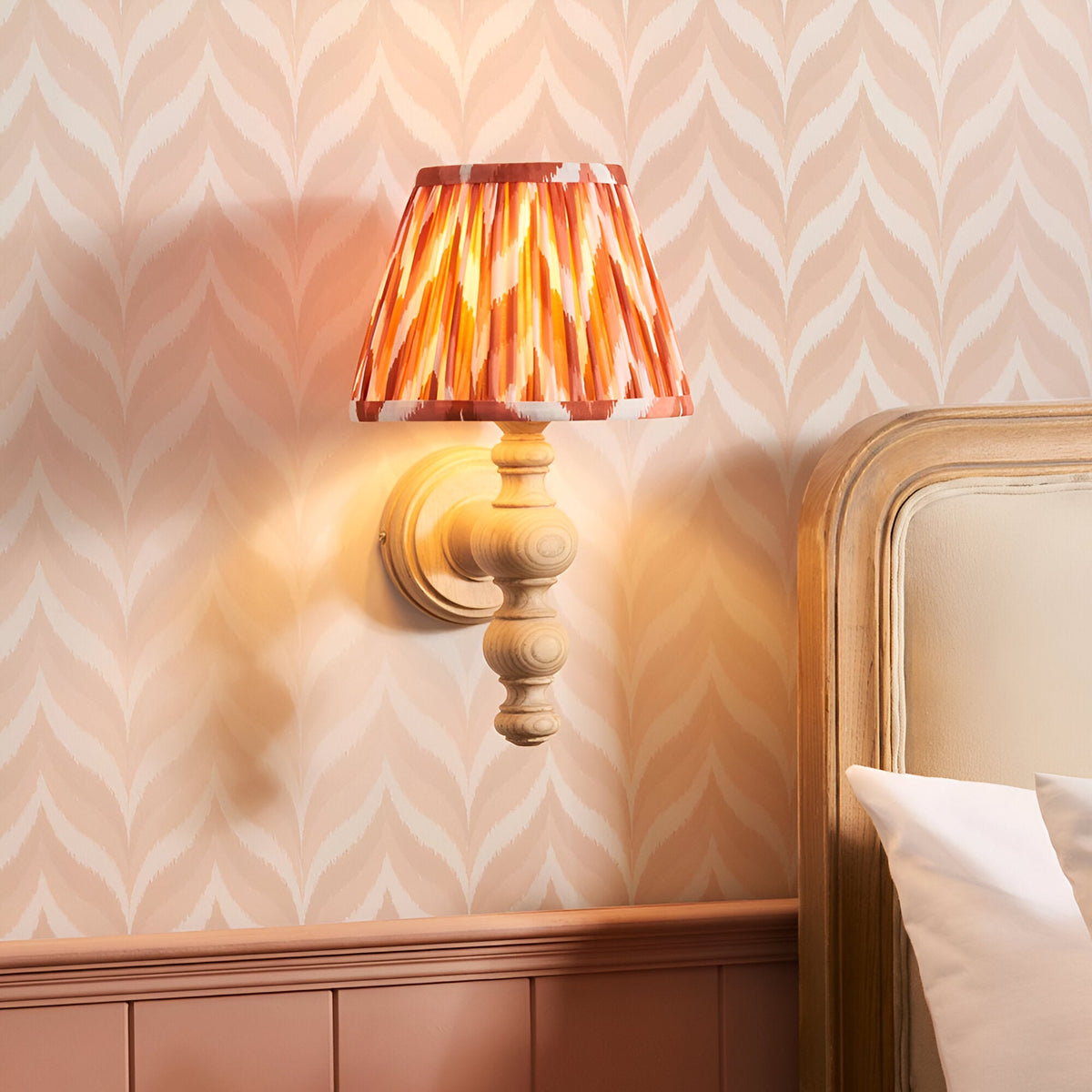 Aldworth Zigzag Wall Light in natural ash wood with turned spindle base and pleated apricot orange patterned shade, shown lit beside a bed headboard.