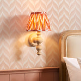 Aldworth Zigzag Wall Light in natural ash wood with turned spindle base and pleated apricot orange patterned shade, shown lit beside a bed headboard.