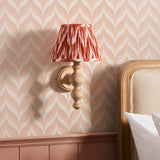 Aldworth Zigzag Wall Light in ash wood with turned spindle base and pleated apricot zigzag shade, mounted beside a bed on pink patterned wallpaper.