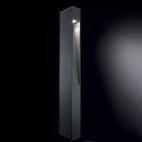 A tall, sleek Nest Outdoor Bollard Light in grey, crafted from powder-coated aluminum, stands illuminated against a dark background. Its minimalist design features clean lines with light softly glowing through Pyrex glass from an angled cut near the top.
