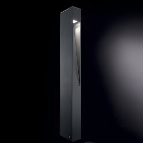 A tall, sleek Nest Outdoor Bollard Light in grey, crafted from powder-coated aluminum, stands illuminated against a dark background. Its minimalist design features clean lines with light softly glowing through Pyrex glass from an angled cut near the top.