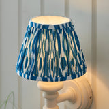 Bevbury Ikat wall light with a pleated blue and white patterned shade on a white turned spindle base, shown illuminated against a panelled wall.