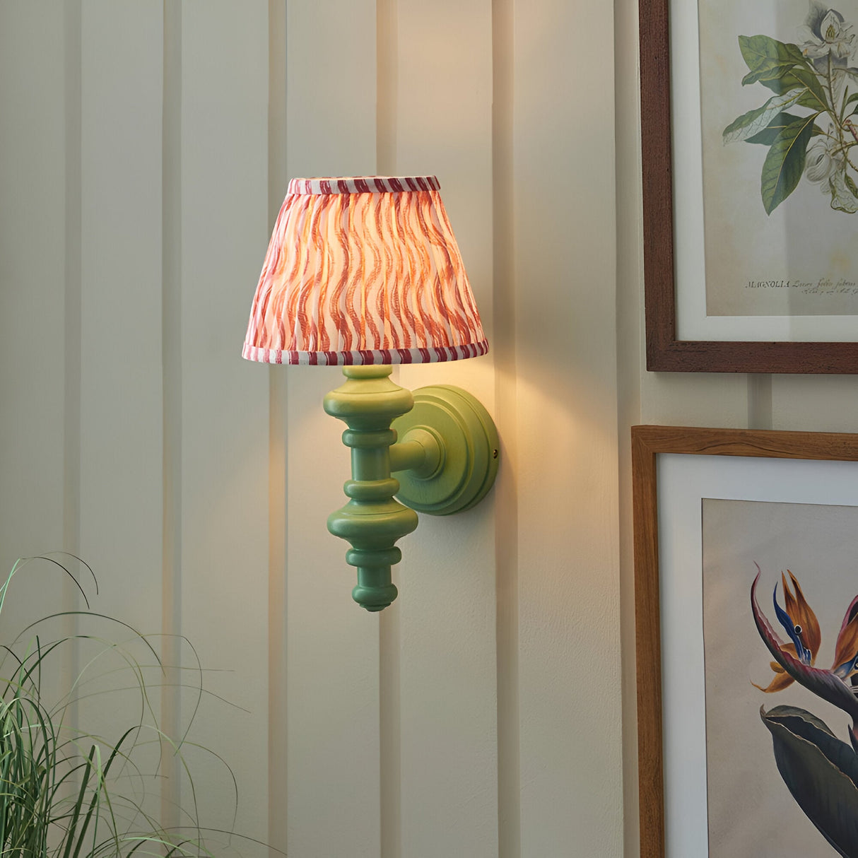 Endon Bevbury Ripple wall light with green turned wooden spindle, brass B22 holder and pink rippled faux linen shade, on panelled wall by botanical prints.