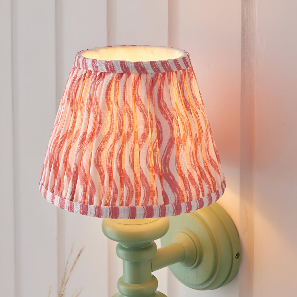Endon Bevbury Ripple wall light with green turned wood spindle, brass lamp holder and tapered pink ripple-print pleated fabric shade, illuminated.