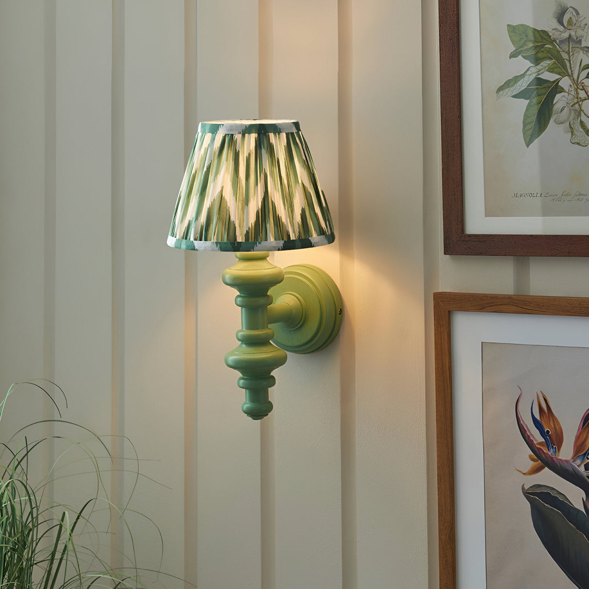 Endon Bevbury Zigzag wall light with green turned wooden spindle and pleated green zigzag faux silk shade, mounted on a panelled wall beside botanical prints.