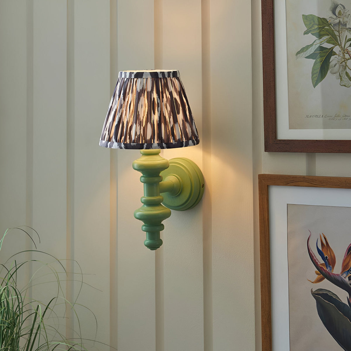 Bevbury Ikat wall light with green turned spindle base, brass mount, and pleated grey-patterned fabric shade, shown mounted beside framed botanical prints.