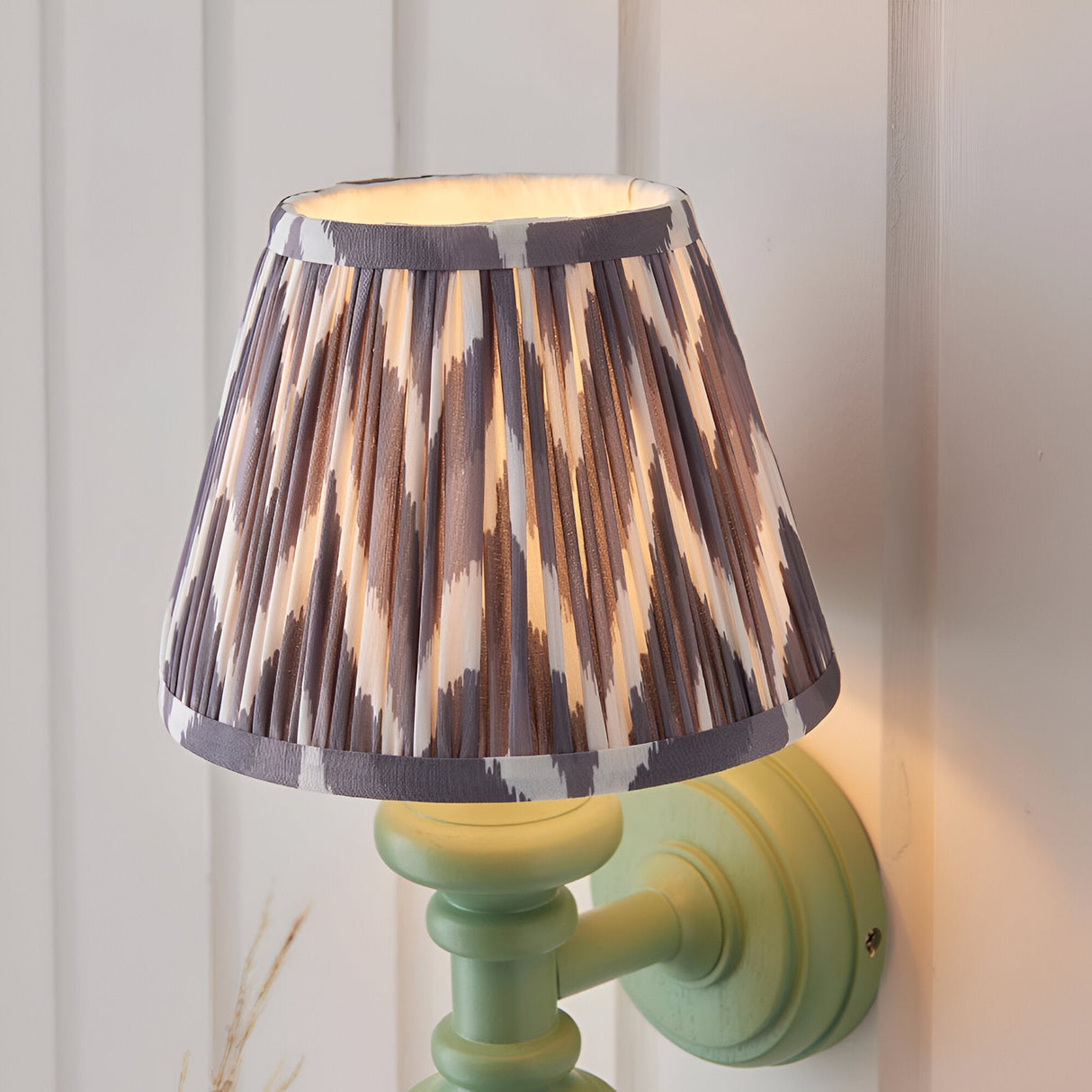 Bevbury Ikat wall light with pleated grey and cream patterned shade on a green turned spindle base, mounted on a panelled wall.