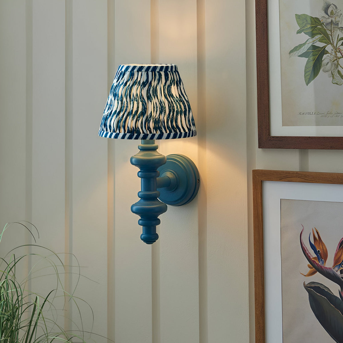 Endon Bevbury Ripple wall light with turned blue wooden spindle and blue ripple-printed tapered fabric shade, mounted on panelled wall beside botanical prints.
