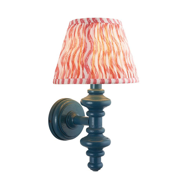 Endon Bevbury Ripple wall light with a blue turned wooden spindle, brass B22 lamp holder and tapered pink rippled faux linen shade on white.