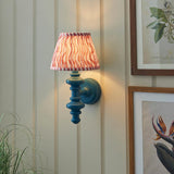 Endon Bevbury Ripple wall light with blue turned wood spindle, brass B22 holder and tapered pink ripple-print faux linen shade, on panelled wall.