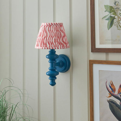 Endon Bevbury Ripple wall light with blue turned wood spindle, brass B22 holder and pink striped faux linen shade, mounted beside framed botanical prints.