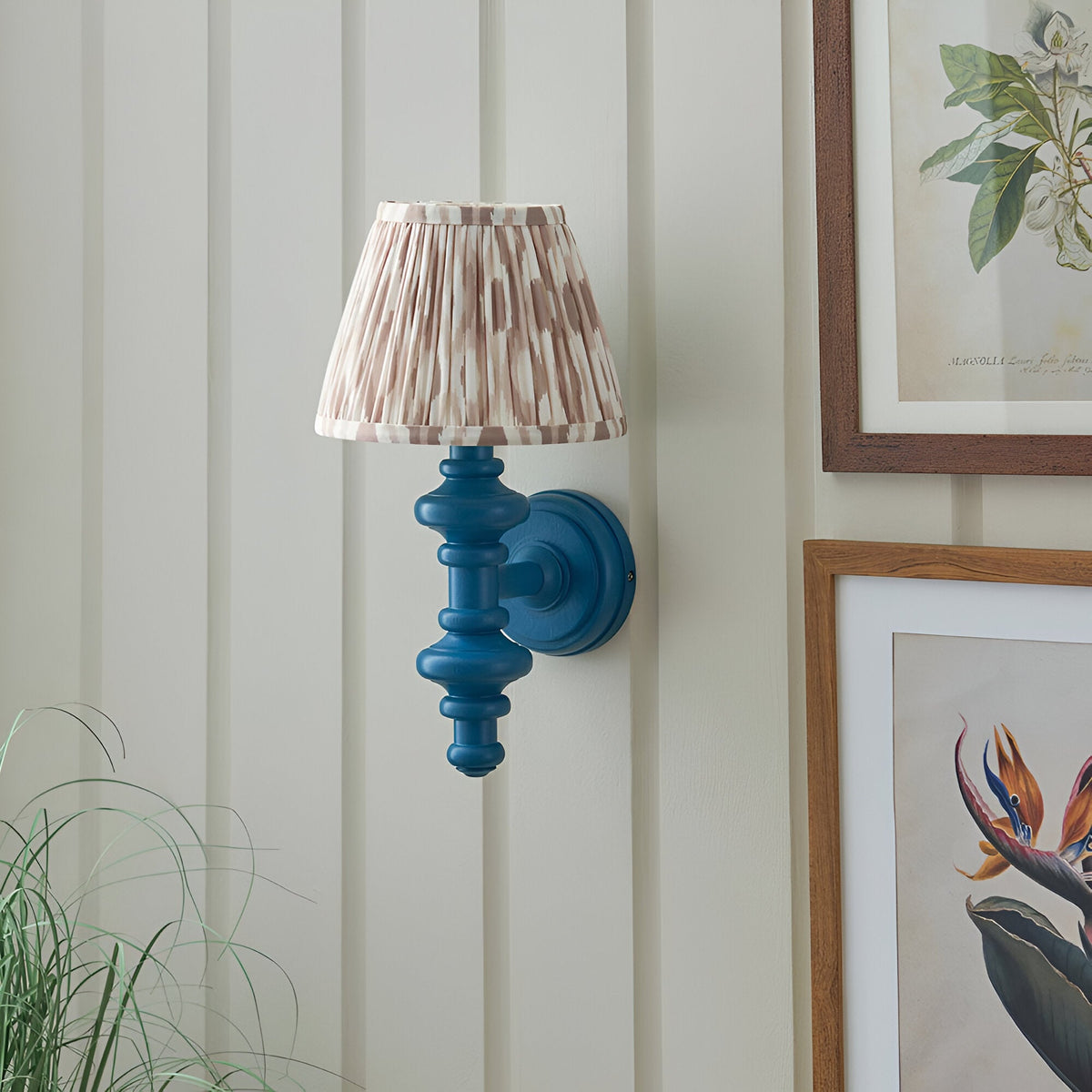 Endon Bevbury Ikat wall light with blue turned wooden spindle and pleated brown ikat faux silk shade, mounted on panelled wall beside botanical prints.