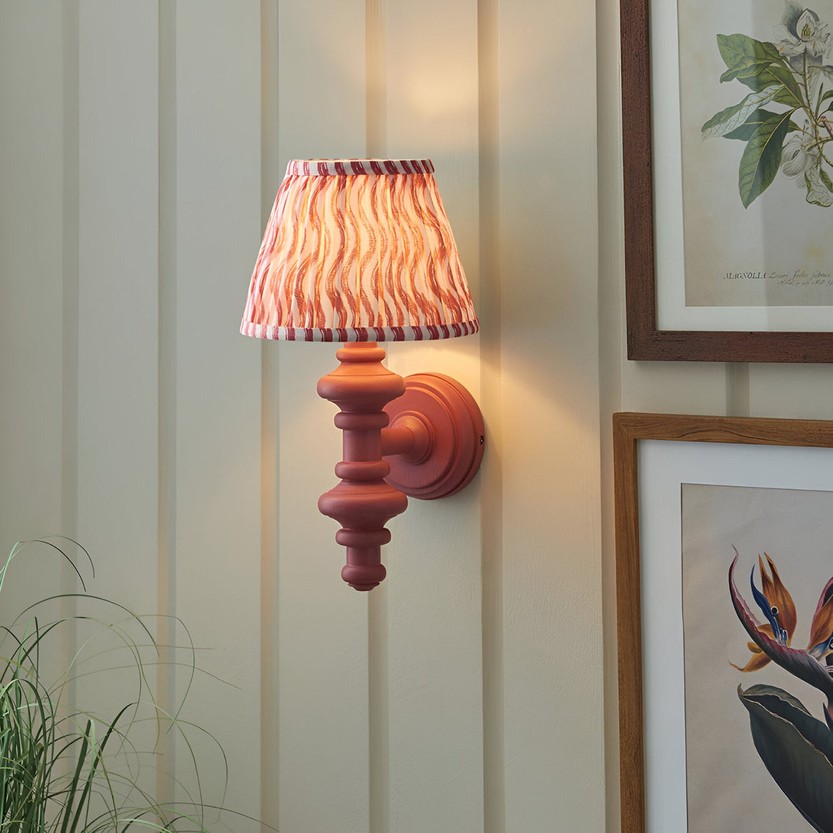 Endon Bevbury Ripple wall light in pink clay with turned wooden spindle and tapered rippled fabric shade, mounted on panelled wall beside botanical prints.