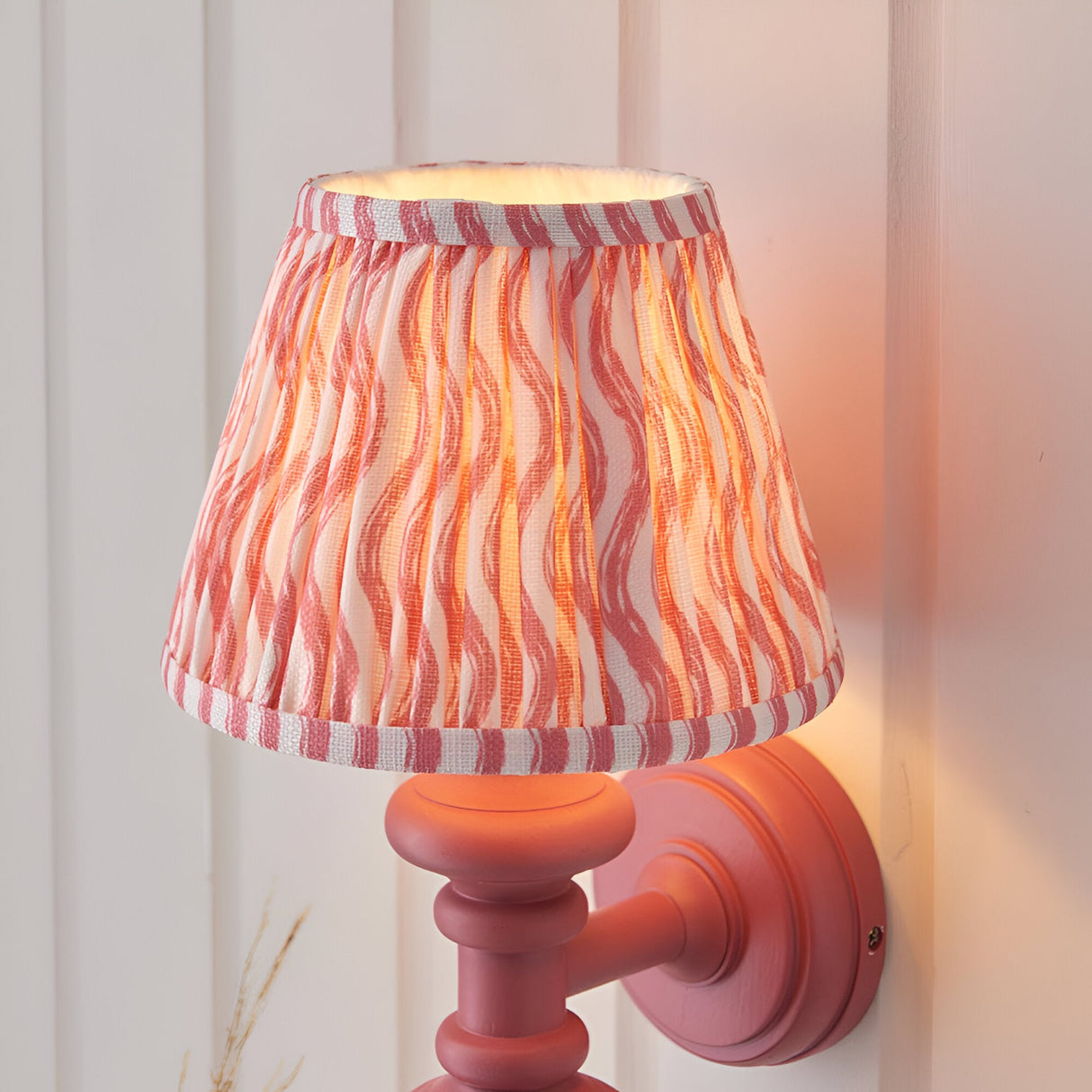 Endon Bevbury Ripple wall light in pink clay with wooden turned spindle and tapered pleated shade in pink rippled faux linen, mounted on panelled wall.