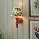 Endon Bevbury Zigzag wall light with pink clay turned wood spindle and green ikat zigzag pleated shade, mounted on a panelled wall beside botanical prints.