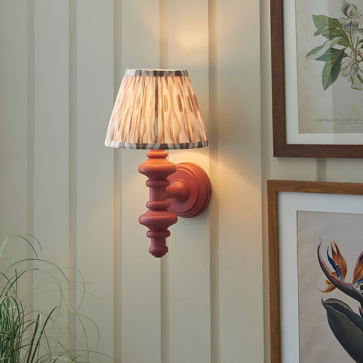 Bevbury Ikat wall light with turned wooden spindle in pink and a pleated Ikat fabric shade, mounted on a panelled wall beside framed botanical prints.