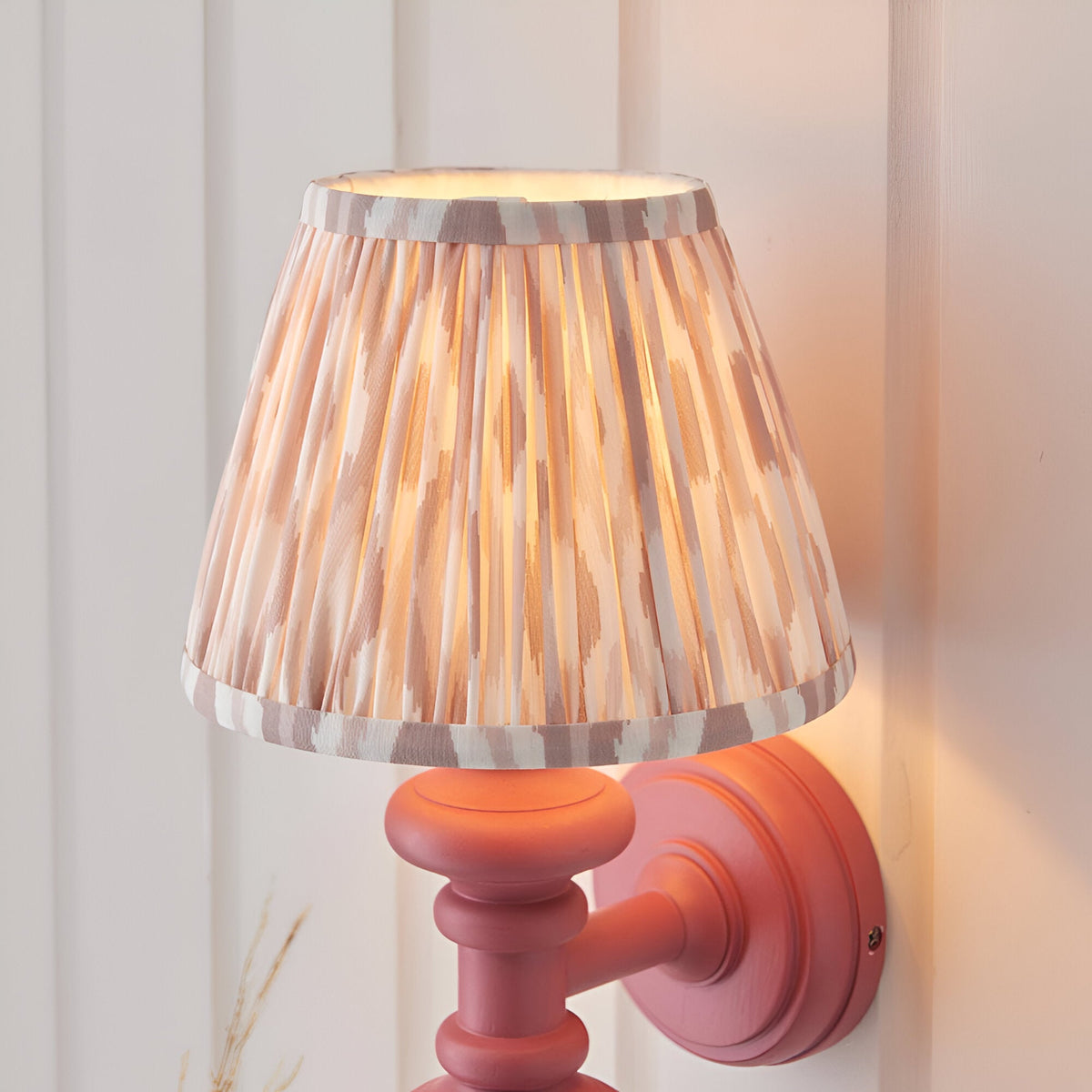 Bevbury Ikat wall light with pink turned wooden spindle base and pleated natural Ikat fabric shade, mounted on a light panelled wall.