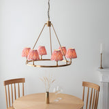 Bevford Ikat 6 Light Pendant in Coral, a circular aged brass hoop chandelier with pleated coral pink fabric shades, shown above a dining table.