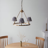 Bevford three-light hoop pendant in aged brass with pleated grey Zigzag fabric shades, shown hanging above a dining table in a neutral room.