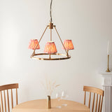 Bevford Ikat 3-Light Pendant in Coral, an aged brass hoop chandelier with three pleated coral pink Ikat fabric shades, hanging above a wooden dining table.