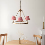 Bevford Ikat 3 Light Pendant in aged brass with coral pink pleated fabric shades on a circular hoop frame, shown above a dining table.