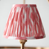 Close-up of a Bevford Ikat 3 Light Pendant shade in pleated coral pink and white fabric, showing a brass gathered trim against a plain light background.