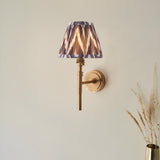 Endon Bevford wall light with aged brass hoop stem and pleated grey zigzag faux silk shade, mounted on a neutral wall beside dried grasses.